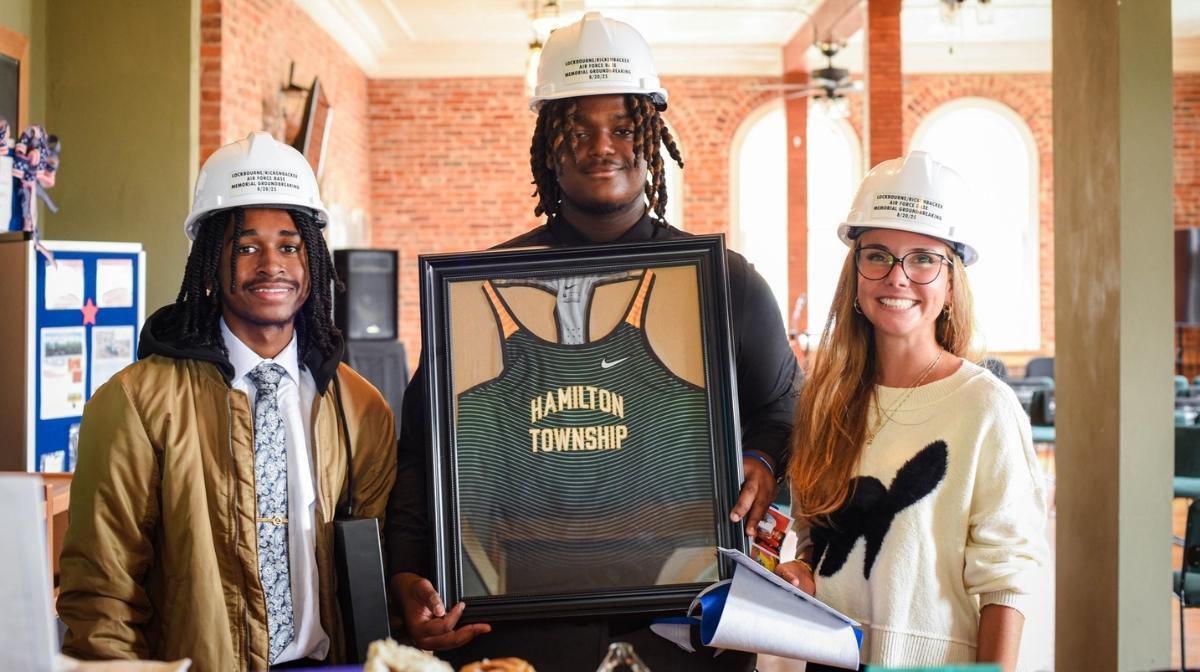 Three people wearing hard hats holding a framed Hamilton Township jersey during a memorial wall ground-breaking event